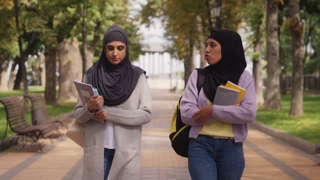 Two multiethnic female students in hijabs communicating while walking in park