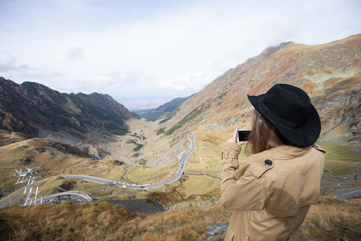 a girl in a raincoat and a black hat takes a photo of a panorama in the mountains on her phone. the girl turns her back to the camera and films the serpentine road on her phone.