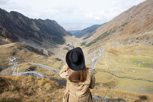 a girl with loose hair and a black hat stands with her back to the camera and looks at the serpentine road in the mountains. Transfagaras, Romania