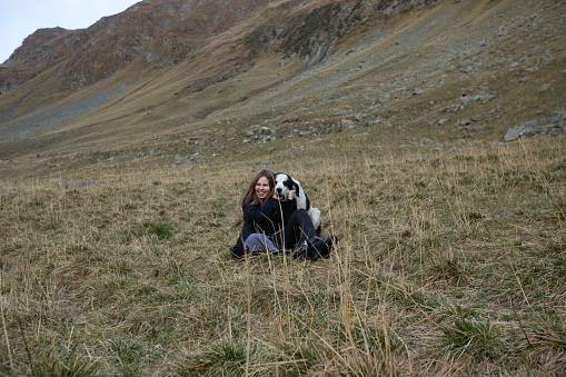 a big dog hugs a girl from behind. a woman plays with a dog in the mountains. the dog lunged at the girl sitting on the grass and laughing. general plan