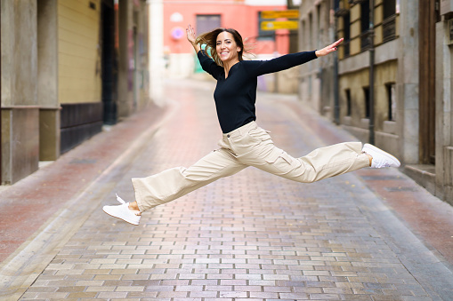 Side view of delighted young female with brown hair in casual clothes and sneakers smiling and looking at camera while jumping on narrow paved street