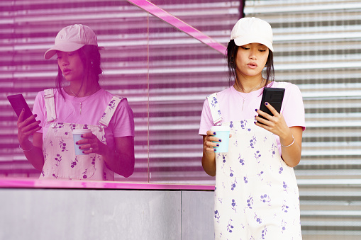Stylish young Asian woman with cup of coffee to go reading news on cellphone while standing near reflective pink wall of modern building in daytime