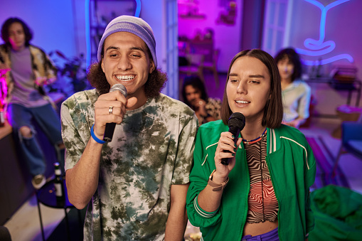 Young multicultural couple with microphones singing karaoke songs in front of tv set screen on background of their friends at home party
