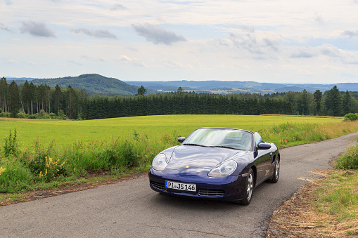 Gerolstein, Germany - June 28, 2021: Blue roadster Porsche Boxster 986 with Eifel mountain panorama. The car is a mid-engine two-seater sports car manufactured by German automobile company Porsche.