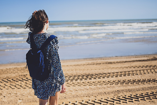 Happy woman walks to sea on beach with blue backpack.