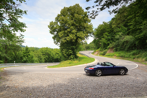 Hunsrück, Germany - June 26, 2021: Blue roadster Porsche Boxster 986 at street with hairpin turn in Hunsrück. The car is a mid-engine two-seater sports car manufactured by German automobile company Porsche.