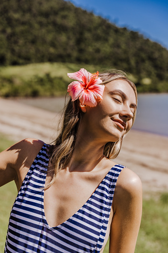 Close up of a lady with a hibiscus flower in her hair and striped swimwear, with her eyes closed and head turned to soak up the sunlight.