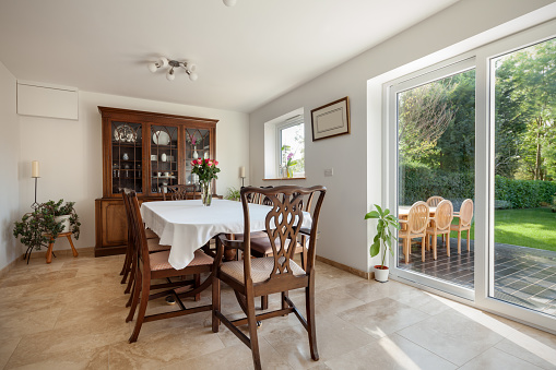 Sunlit dining room within modern house with traditional wooden furniture and sideboard, patio doors overlooking garden and white painted walls
