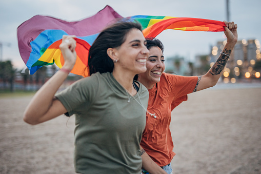 Two women, lesbian couple holding and running with rainbow flag together outdoors on the beach.