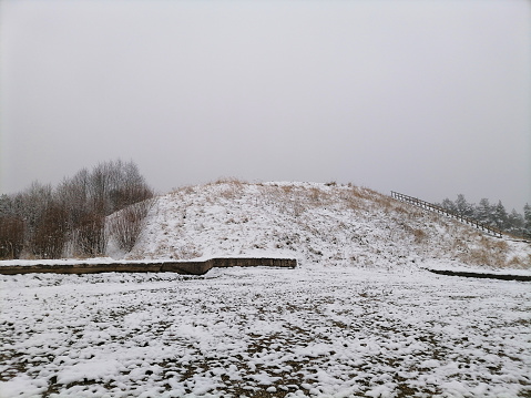 Hill in countryside. Snow on it. Stairs leading to the top of it. Cloudy day with white and gray clouds in sky. Winter season. Nature. Salduves piliakalnis.