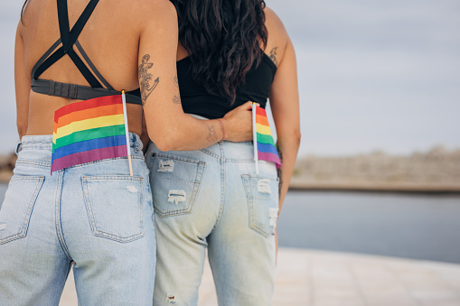 Two women, lesbian couple with rainbow flags together outdoors by the sea.