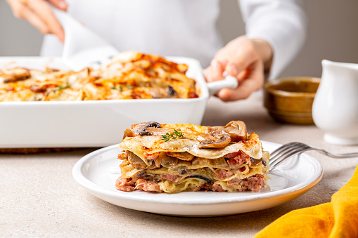 Serving, cooking homemade white lasagna with porcini and champignon mushrooms, onion and meat sausages. Pasta with parmesan cheese and bechamel sauce. Woman hands holding casserole.