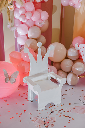 Arch decorated pink balloons, confetti, paper decor butterfly, and wooden white chair. Children's photo zone. Birthday party for girl on a background photo wall. Closeup.