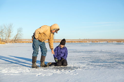 Girl sitting on the ice while ice fishing. She is waiting patiently for a fish to bite, as Grandpa gives her tips on what to do.