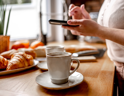 Girl hand stirs coffee cappuccino in cup with spoon and looking smartphone screen. Woman with creamy espresso and mobile phone at kitchen