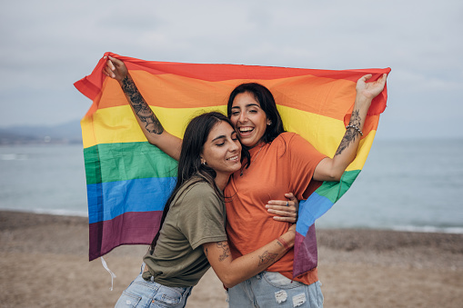 Two women, lesbian couple holding a rainbow flag together outdoors on the beach by the sea.
