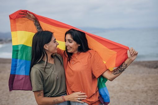 Two women, lesbian couple holding a rainbow flag together outdoors on the beach by the sea.
