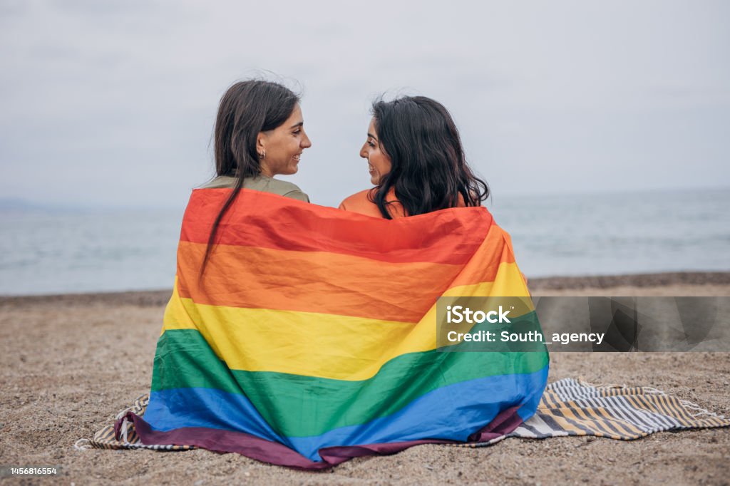 Lesbian couple on the beach Two women, lesbian couple covered in rainbow flag sitting together outdoors on the beach. LGBTQIA People Stock Photo
