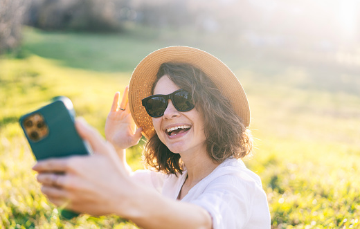 Young pretty woman in sunglasses and straw hat taking selfie on smartphone in green field enjoying sunny summer