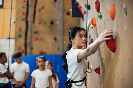 A shot of a teen schoolgirl gripping hand and foot holds as she scales a climbing wall during her PE class in a modern indoor facility in Gateshead, England.