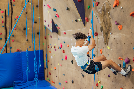 A side-view shot of a teen schoolboy using hand and foot holds as he scales a climbing wall during his PE class in a modern indoor facility in Gateshead, England.