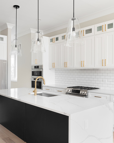 A luxury white kitchen with black pendant lights hanging above a waterfall granite island, stainless steel appliances, and gold hardware and faucet.