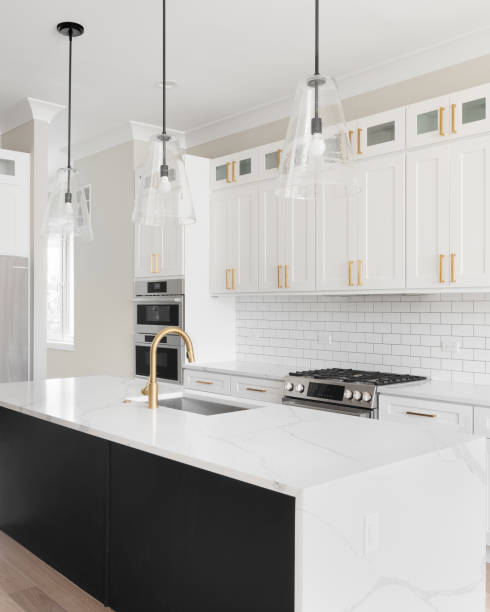 A white and black kitchen with lights hanging above the waterfall granite island. A luxury white kitchen with black pendant lights hanging above a waterfall granite island, stainless steel appliances, and gold hardware and faucet. quartz stock pictures, royalty-free photos & images