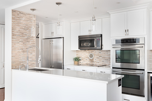 A small renovated, white kitchen with a rock tile backsplash, glass pendant lights hanging above the island, stainless steel appliances, and a granite countertop.