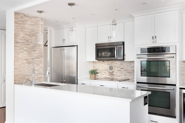 A kitchen with stainless steel appliances and tiled backsplash. A small renovated, white kitchen with a rock tile backsplash, glass pendant lights hanging above the island, stainless steel appliances, and a granite countertop. hanging stone stock pictures, royalty-free photos & images