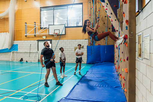 A side view shot of a teen schoolgirl using hand and foot holds as she scales a climbing wall during her PE class in a modern indoor facility in Gateshead, England. A climbing instructor and her friends are with her.