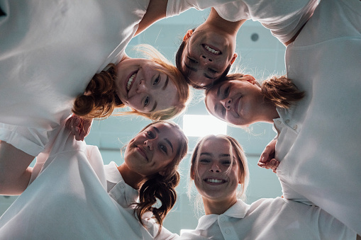 A directly below shot of a group of schoolgirl friends wearing school uniforms, standing in a huddle with their heads together, they are smiling and looking at the camera.