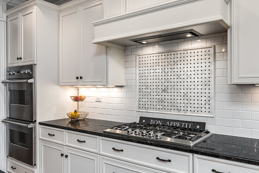 A luxury white kitchen with a custom tiled backsplash above the stovetop, under cabinet lighting, and black granite counter tops.