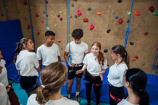 A shot of a small group of teen schoolboys and schoolgirls preparing to scale a climbing wall, they are standing together near the climbing wall during their PE class in a modern indoor facility in Gateshead, England.