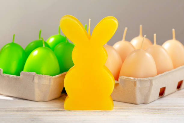 Easter.A yellow candle in the shape of a rabbit stands on the white wooden table. Behind him are green and beige egg-shaped candles in trays