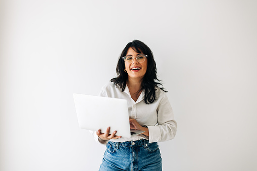 Business woman smiling at the camera while holding a laptop in an office. Professional woman standing against an office wall dressed in business casual.