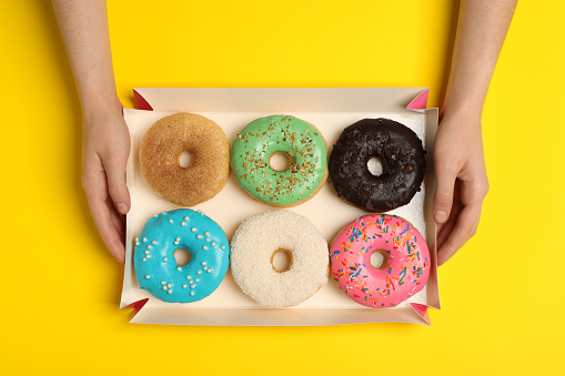 Woman with box of delicious donuts on yellow background, top view