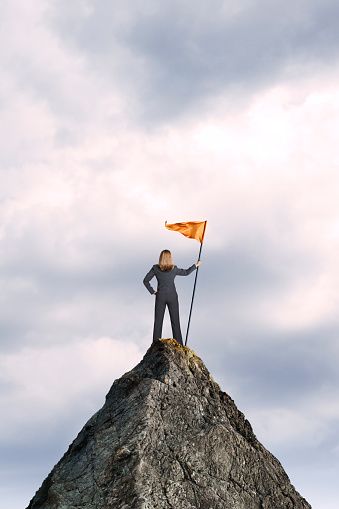 A businesswoman stands at the top of a mountain peak as she holds a large orange flag attached to a pole. She stands with her back to the camera as she looks out into the distance.