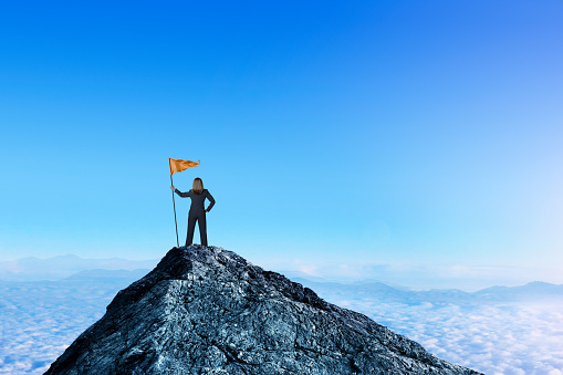 A businesswoman stands at the top of a mountain peak as she holds a large orange flag attached to a pole. She stands with her back to the camera as she looks out into the distance toward a distant mountain range that sits on the horizon.