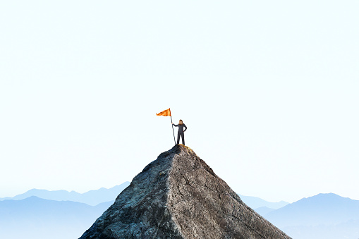 A businesswoman stands at the top of a mountain peak as she holds a large orange flag attached to a pole. She stands with her back to the camera as she looks out into the distance toward a distant mountain range that sits on the horizon.