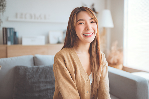 Confident smiling young adult asian woman looking at camera while sitting at home, Happy beautiful lady pretty face dental smile posing alone indoors, Slow motion close up view portrait