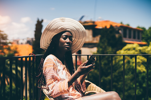 A shot of a young sensual woman with a wide-brimmed hat and long colorful braids, wearing a colorful oversized shirt, texting with both hands while soaking up some sun at the apartment balcony