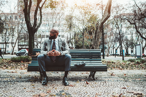 An elegant executive unshaven but hairless man of African descent in a suit sitting on the extremity of a dark green wooden street bench with his wool beret next to him in winter scenery