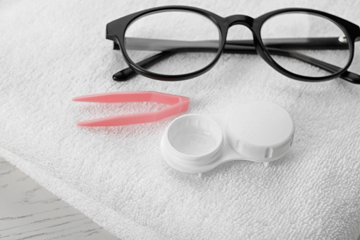 Case with contact lenses, tweezers, glasses and towel on white table, closeup