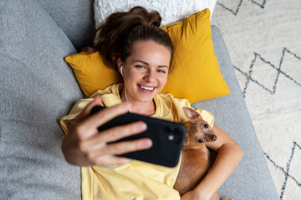 istock Young woman taking selfie using her smartphone while lying down on sofa with little dog. 1456838363