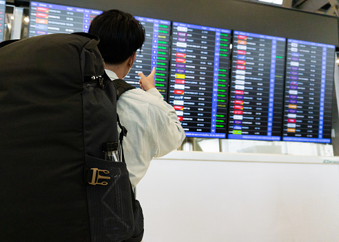 Young backpacker man looking at departure boarding in the airport  while waiting to his plane for traveling alone.