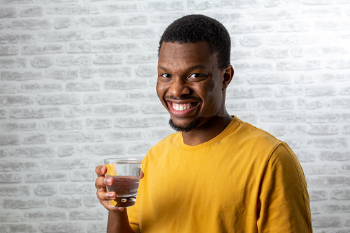 Young handsome black man drinking glass of water smiling happy with open mouth. smiling friendly gesturing excellent symbol. Young man holding a glass of water on white background