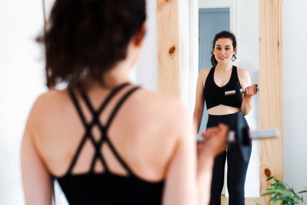 istock Young woman looking in the mirror while lifting dumbbel 1456849619
