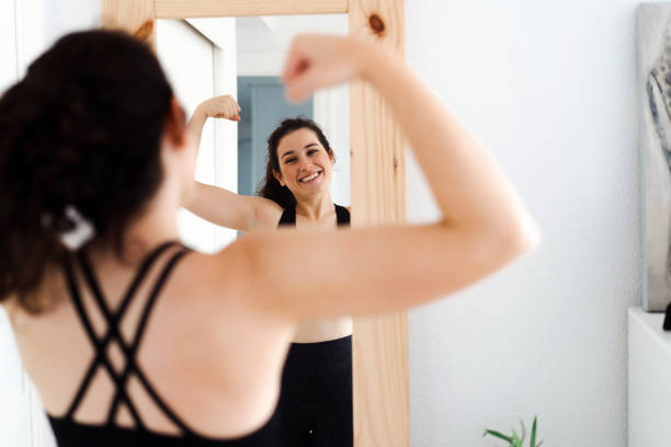 Funny young woman flexing arms looking in the mirror after training - fotografia de stock