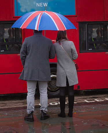 Back view of well-dressed young couple with British flag umbrella waiting in front af a red London double-decker bus. London, England - December 31, 2022.