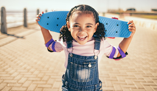 Child, skateboard and excited for fun activity outdoor on promenade with smile, happiness and energy on summer vacation. Portrait of black girl with safety gear for elbow for skating or skateboarding